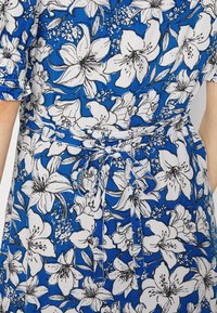 Vestido floral azul feito de um tecido leve, apresentando grandes flores brancas com contornos pretos, uma cintura com cinto e mangas curtas.