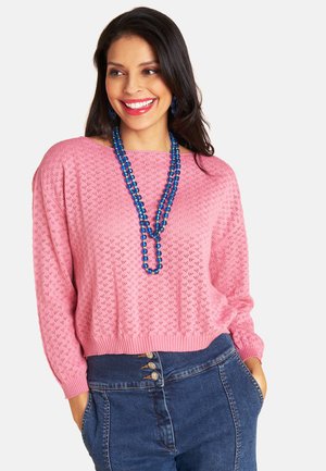 Frau lächelt, trägt einen rosa gestrickten Pullover, eine mehrlagige blaue Perlenkette und eine hoch taillierte blaue Jeans mit Knöpfen, Hände in den Taschen.