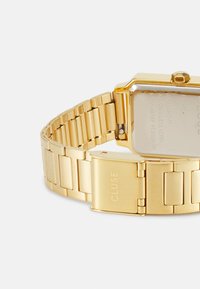 Montre-bracelet en acier inoxydable doré avec un boîtier rectangulaire, un bracelet à maillons et une boucle comportant un logo gravé. Le dos affiche des détails sur le mouvement et la résistance à l'eau.