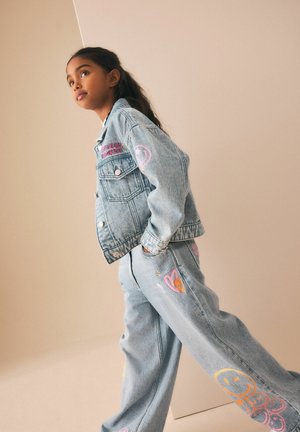 Junge Person geht, trägt eine hellblaue Denimjacke und Hose mit bunten, bestickten Aufnähern an Ärmeln und Beinen vor beigem Hintergrund.