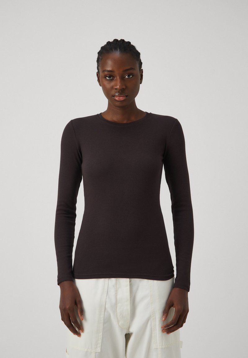 Haut ajusté noir à manches longues en côtes, col rond avec des poignets sans couture. Texture lisse avec une coupe légèrement extensible, associé à un pantalon de couleur claire.