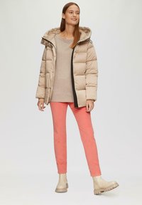 Beige pufferjack met een capuchon, zwarte ritssluiting, lichtbeige sweater, koraalkleurige broek en lichtbruine enkelboots met een dikke zool.