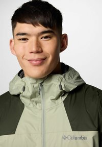 Młody mężczyzna z krótkimi ciemnymi włosami, uśmiechający się, ubrany w jasnoszara i oliwkową kurtkę Columbia z zamkiem błyskawicznym przy kapture.