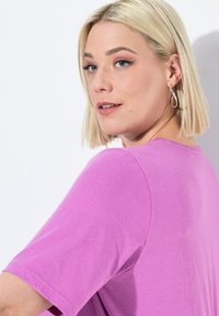 Tričko s krátkým rukávem v jasně růžové barvě, vyrobené z bavlny, s kulatým výstřihem a hladkým povrchem. Modelka má bob účes a nosí stříbrné kruhové náušnice.