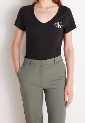 Femme portant un t-shirt noir Calvin Klein Jeans à col en V, rentré dans un pantalon taille haute vert olive, les mains dans les poches.