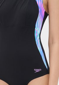 Maillot de bain noir avec un design structuré et des panneaux latéraux colorés. Le matériau est lisse et extensible ; il comprend un logo Speedo rose en bas à gauche.