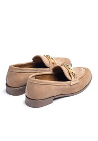 Tan suède loafers met een lage bruine hak, voorzien van een opvallende goudkleurige gesp aan de voorkant en gestikte accenten langs de randen.