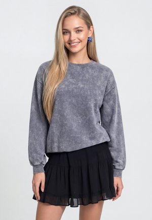 Jonge vrouw met lang blond haar die een gewassen grijze trui, een zwarte gelaagde rok en blauwe oorbellen in stervorm draagt, glimlachend naar de camera.