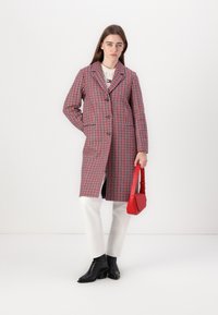 Een rood en marineblauw houndstooth-jas met een geknipte kraag, enkele knoop, zijzakken, gecombineerd met witte jeans en zwarte enkellaarsjes, terwijl ze een rode tas vasthoudt.