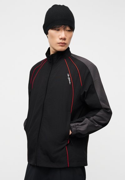 Νέος άνδρας που φοράει μαύρο σκουφί και μαύρο μπουφάν με φερμουάρ, με γκρι μανίκια και κόκκινη διακοσμητική ταινία, κοιτάζοντας προς τα κάτω αριστερά με τα χέρια στις τσέπες.