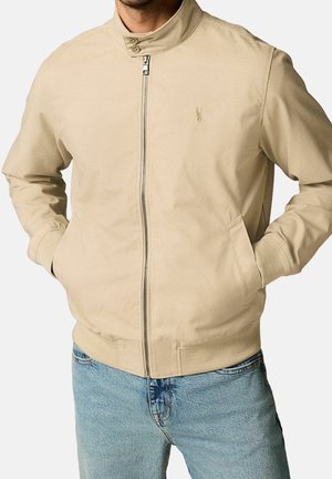 Mand iført en beige lynlåstrøje med ståkrave og lyseblå jeans, hænderne i jakke-lommerne, foran en ensfarvet baggrund.