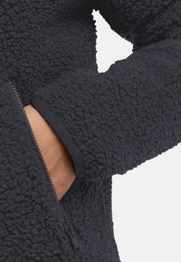 Fleece jas in donkergrijs met een gestructureerd oppervlak, voorzien van een zijzak met rits en elastische manchetten voor een comfortabele pasvorm.