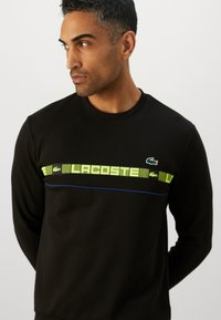 Svart hettegenser med rund hals, med et neongrønt grafisk bånd over brystet og en blå linje under. Lacoste-logo på venstre side.