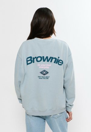 Persona con cabello largo y oscuro que lleva una sudadera gris claro con la palabra "Brownie" y texto motivacional impresos en la parte trasera, combinada con jeans azul claro.