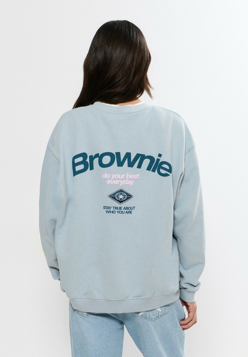 Persona con cabello largo y oscuro que lleva una sudadera gris claro con la palabra "Brownie" y texto motivacional impresos en la parte trasera, combinada con jeans azul claro.