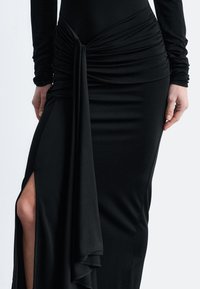 Schwarzes Kleid aus glattem Stoff, mit einer gerafften Gestaltung in der Taille, einem seitlichen Schlitz und langen Ärmeln mit gerafften Manschetten.