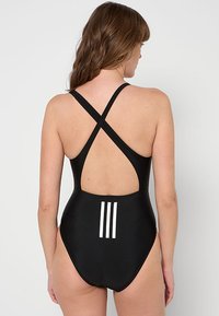 Costume da bagno intero nero con spalline incrociate, schiena scoperta e tre strisce bianche nella parte bassa della schiena. Tessuto dalla consistenza liscia.