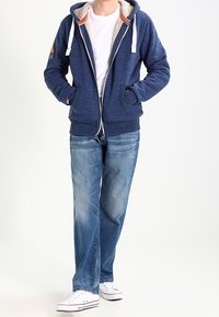 Námořnická mikina na zip s bílými šňůrkami a šedým chlupatým podšívkou, kombinovaná s světle modrými džíny a bílými teniskami. Neformální střih a styl.