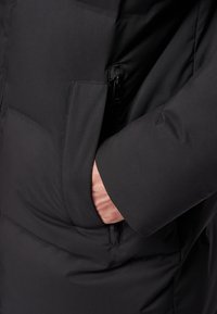 Giacca isolata nera con tasca con zip e una texture di tessuto liscia; primo piano della manica con la mano posizionata nella tasca.