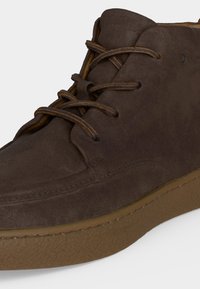 Baskets montantes en daim marron avec lacets assortis et semelle en caoutchouc brun texturée, montrées en gros plan sur fond blanc.