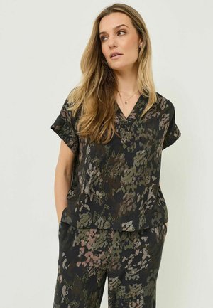 Donna con lunghi capelli biondi che indossa una blusa a maniche corte con fantasia floreale scura e pantaloni abbinati, in piedi con le mani nelle tasche, che guarda di lato.