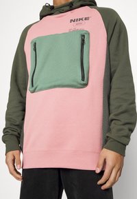 Man som bär en rosa och grön Nike-hoodie med en grön dragkedjeficka fram och ärmar i olivgrön, kombinerat med svarta byxor.
