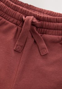 Pantalon de survêtement bordeaux avec une taille élastique équipée d'un cordon de serrage, une texture lisse et un détail de couture subtile près de la taille.
