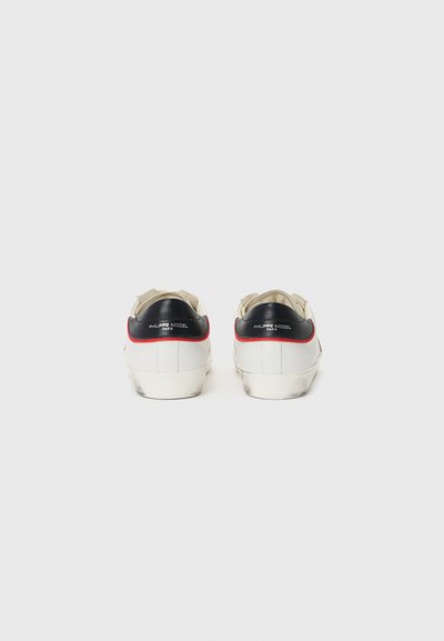 Baskets blanches avec une finition texturée, des accents noirs au niveau du talon et des bordures rouges. Design minimaliste avec un aspect légèrement usé sur les semelles.
