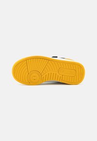 Hvit sneaker med ytre såle av teksturert gult gummi, utstyrt med dype spor for grep og et sirkulært emblem nær hælen.