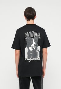 Černé tričko s grafickým motivem psa na zadní straně, s bílým textem "ADIDAS" a "originals" nad a pod obrázkem.