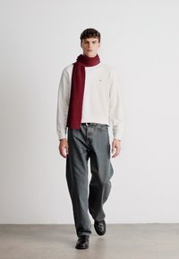 Bílý dlouhý rukáv, bordó šál, volné šedé kalhoty, černé boty. Jednoduchý design s kulatým výstřihem a decentním logem na tričku.