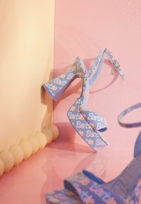 Talons hauts bleus avec un tissu texturé arborant des inscriptions colorées "Barbie" et des cœurs. Bride fine autour de la cheville et design à talon épais.