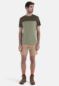 Camicia a maniche corte da uomo con parte superiore verde oliva, corpo verde oliva chiaro, abbinata a pantaloni corti beige e scarponi da trekking marroni. Calzini arancioni visibili.