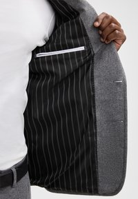 Grau sportliches Sakko mit einem strukturierten Außenmaterial, das ein schwarzes gestreiftes Innenfutter und eine Brusttasche aufweist. Die Revers sind eingekerbt und die Nähte sind sichtbar.