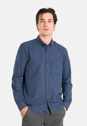 Young man with dark hair wearing a blue button-up shirt and gray pants, standing with hands in pockets against a plain background.