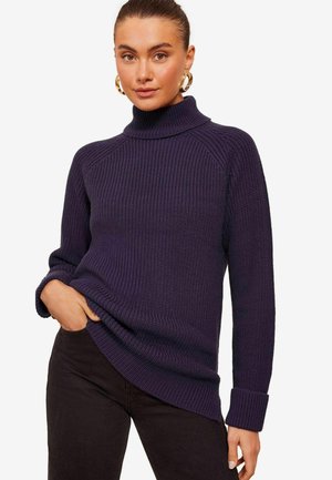 Lila gerippter Pullover mit hohem Rollkragen, ausgestattet mit langen Ärmeln und umgeschlagenen Bündchen, kombiniert mit dunklen Hosen. Sichtbares Detail des strukturierten Stoffs.