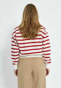 Maglione a righe rosse e bianche con maniche lunghe e polsini a coste, abbinato a pantaloni beige a gamba larga. Focus sul motivo a righe orizzontali.