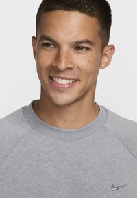 Szary sweatshirt z okrągłym dekoltem i rękawami raglanowymi. Posiada małe czarne logo Nike na klatce piersiowej. Gładka, prosta faktura materiału.