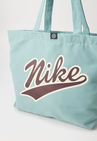 Bolsa de lona azul claro con un gran logo curvado en marrón oscuro de "Nike" en la parte frontal, con dobles asas para el hombro.