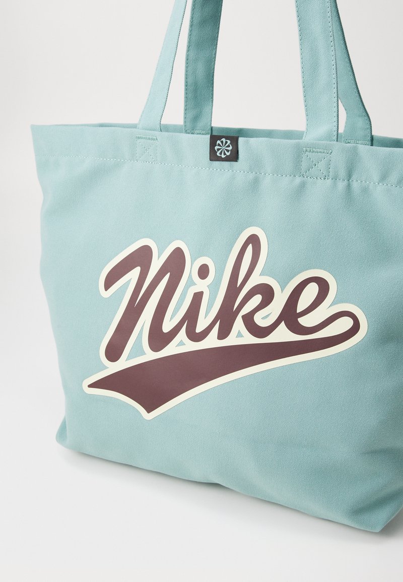 Bolsa de lona azul claro con un gran logo curvado en marrón oscuro de "Nike" en la parte frontal, con dobles asas para el hombro.