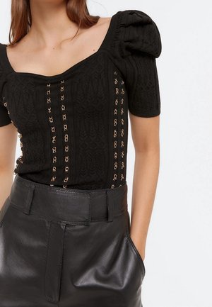 Vrouw draagt een zwart gebreid shirt met opgeblazen korte mouwen en metalen kettingdetails, gecombineerd met een zwart leren broek met hoge taille en handen in de zakken.
