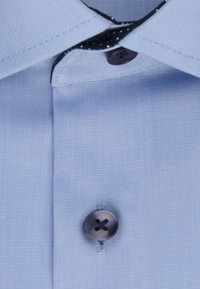 Camicetta azzurro chiaro con tessuto texture, colletto a punta e bottone scuro con quattro fori. Rifinita con un colletto interno a fantasia.