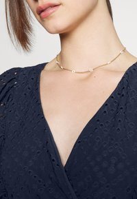 Kultaketju, jossa on pieniä kukkaominaisuuksia ja hentoa ketjua, joka on käytössä kaulassa tummansinisessä tekstuuripinnassa.