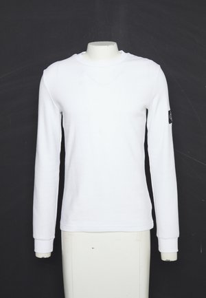 Chemise blanche à manches longues et col rond sur un mannequin sans tête avec un petit patch logo noir sur la manche gauche sur fond noir.