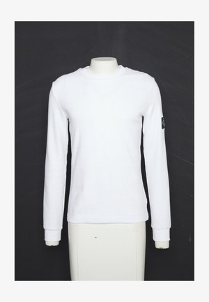 Chemise blanche à manches longues et col rond sur un mannequin sans tête avec un petit patch logo noir sur la manche gauche sur fond noir.
