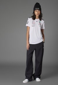 Camisa blanca de manga corta con detalles y logotipos negros, combinada con pantalones anchos negros. Los accesorios incluyen un gorro negro y zapatillas blancas.