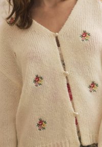 Cardigan lavorato a maglia color crema con scollo a v, ricami floreali e chiusura con bottone perla. Tessuto morbido e vestibilità ampia con dettagli decorativi.