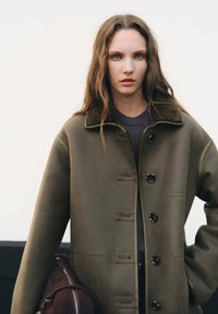 Cappotto di pelle verde oliva con colletto, grandi bottoni e tasche laterali. La modella tiene una borsa marrone, mettendo in risalto la vestibilità rilassata e la texture del cappotto.