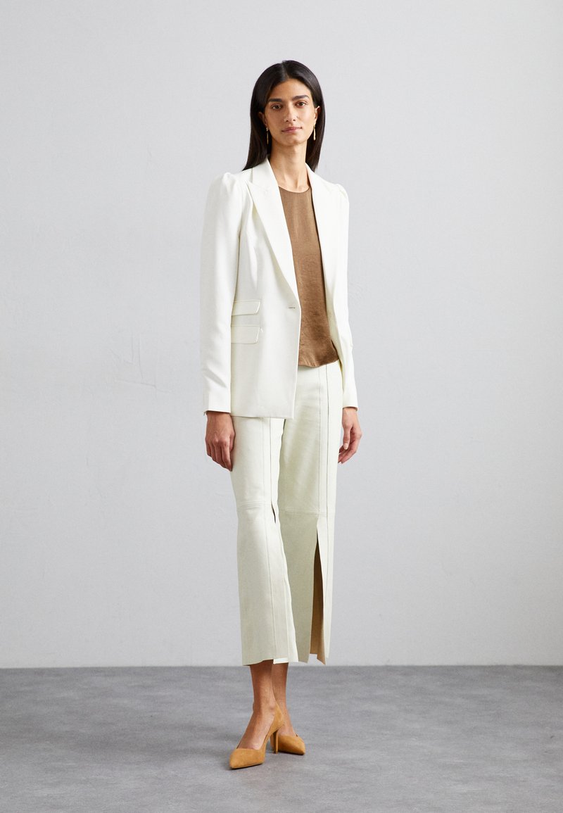 Blazer blanc à épaules structurées, associé à un haut marron et un pantalon crème avec fentes à l'avant, complété par des talons beige.