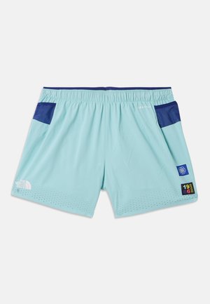 Mand stående iført lyseblå træningsshorts og t-shirt, orange kasket og sokker samt hvide løbesko mod en ensfarvet hvid baggrund.